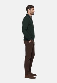 Chaqueta de forro polar verde oscuro para hombre con cremallera, combinada con pantalones marrones y zapatos de slip-on marrones. Presenta un cuello con solapas y bolsillos laterales.