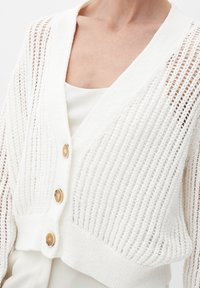 Vit stickad cardigan med öppet vävstruktur, V-ringning och tre gyllene knappar. Kort längd och ribbad band vid nederkanten.