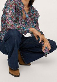 Blusa floral con mangas acampanadas, que presenta flores vibrantes en rosa, azul y verde sobre un fondo negro, combinada con jeans de mezclilla oscura y botas marrones.