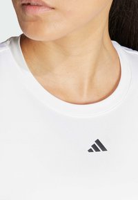 Biała sportowa koszulka z okrągłym dekoltem i krótkimi rękawami, z czarnym logo Adidas w pobliżu kołnierza. Gładki, lekki materiał.