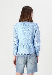 Blusa azzurra chiara con motivo ad occhio ricamato, maniche lunghe, scollatura ruffled e orlo smerlato. Abbinata a pantaloni shorts in denim.