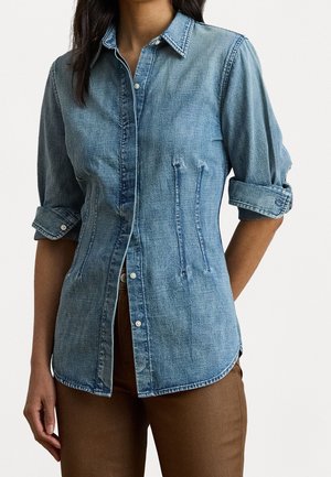 Femme portant une chemise en jean bleu ajustée à manches retroussées et un pantalon marron, debout devant un fond uni.