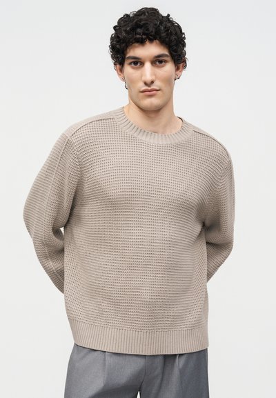 Jeune homme aux cheveux bouclés foncés, portant un pull beige texturé et un pantalon gris, debout devant un fond clair uni.