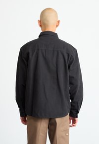 Black cotton shirt with a collar, visible stitching details, and buttoned cuffs. Features a relaxed fit and a straight back design.