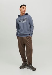 Niebieska bluza z kapturem z logo i napisem, w połączeniu z brązowymi zwężanymi spodniami i szarymi sneakersami. Gładka tekstura, casualowy krój i swobodny styl.