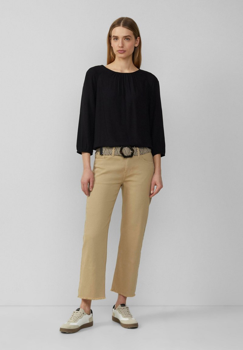 Zwarte, loszittende blouse met gathers bij de hals, lichtbeige wijde broek, een patroonriem en witte sneakers met zwarte accenten.