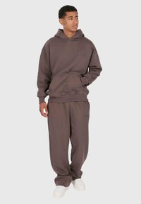 Junger Mann in braunem Hoodie und passenden Hosen steht mit einer Hand in der Tasche, schaut zur Seite und trägt weiße Sneaker.