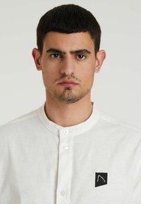 Camicia bianca con colletto realizzata in tessuto leggero, caratterizzata da un piccolo logo ricamato nero sul lato sinistro del petto, con chiusura a bottoni.