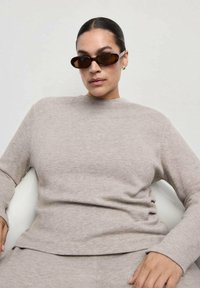 Sweat-shirt beige à manches longues en matière douce, avec un col montant et une coupe décontractée. Porté avec des lunettes de soleil ovales marron.