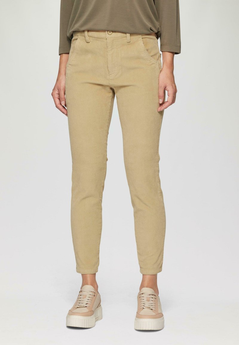 Beige Cordhosen im Slim-Fit, mit frontalen Taschen, Gürtelschlaufen und einem strukturierten Stoff. Kombiniert mit hellen Schuhen.