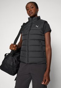 Vestă puffer neagră cu guler înalt, fermoar frontal și logo. Purtată peste un top cu mânecă scurtă, asociată cu o geantă sport neagră cu accente din plasă.