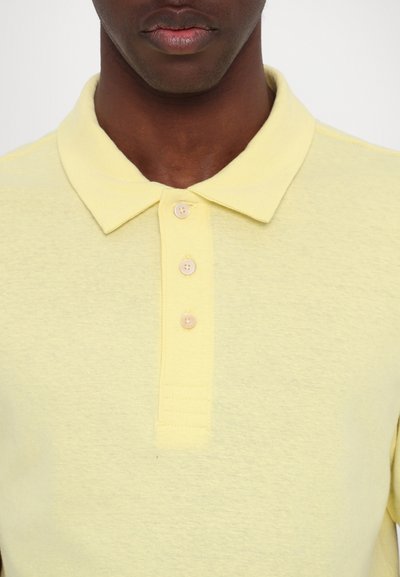 Heledad kollane polo-särk, millel on krae, kolm nööpaukku ja tekstuuriga kangas. Lihtne disain, ilma mustrite või aktsentideta.