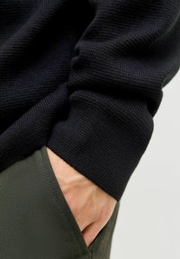 Maglione nero lavorato a maglia con tessuto testurizzato e polsini aderenti. Pantaloni verdi con una tasca laterale, in cui è visibile una mano. Tessuto morbido in entrambi i capi.