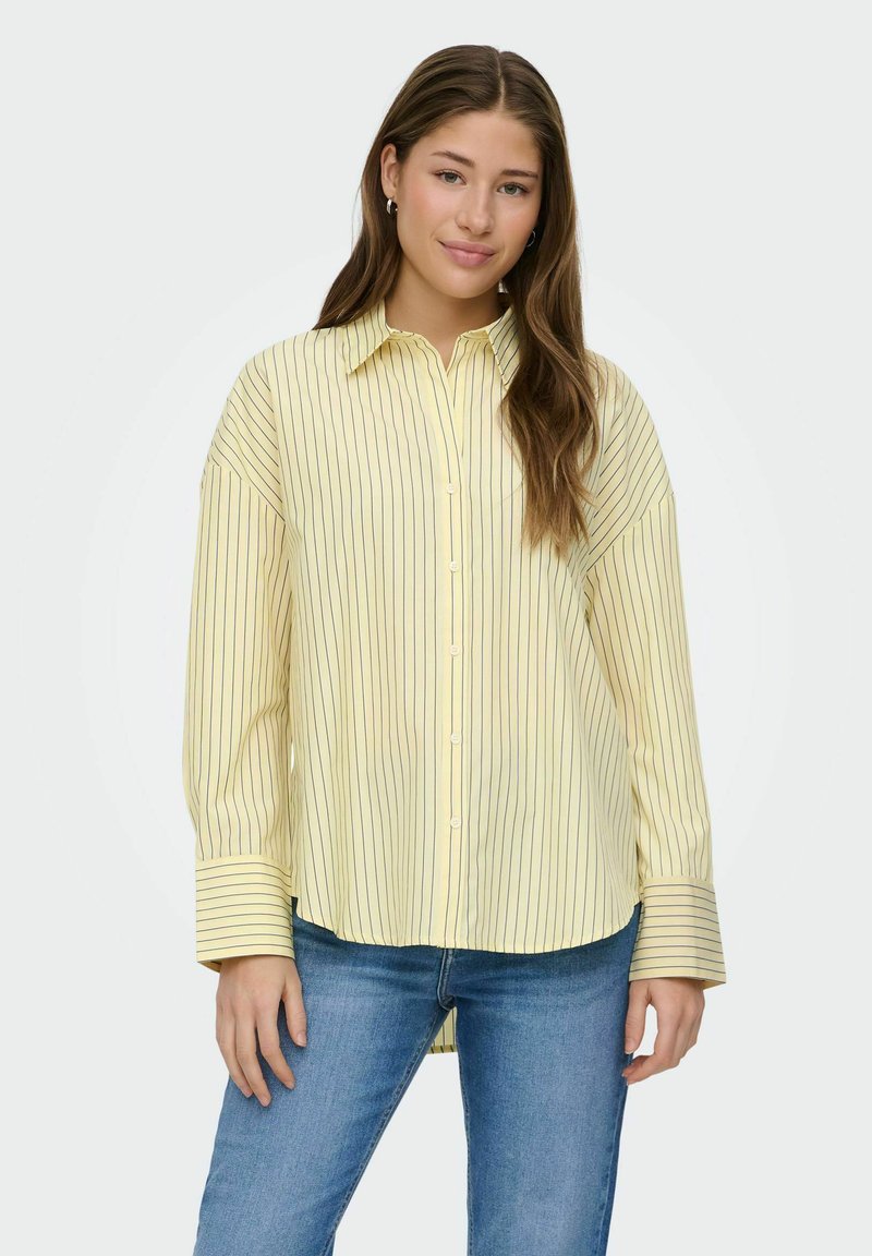 Jeune femme aux longs cheveux bruns portant une chemise jaune ample à boutons avec de fines rayures noires et un jean bleu, se tenant devant un fond blanc.
