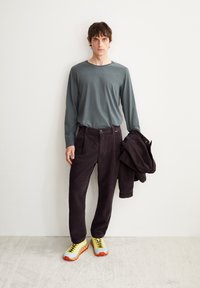 Graues Langarmhemd, dunkle Cordhosen und eine strukturierte schwarze Jacke. Helle gelbe und orange Sneaker mit einem ausgeprägten Sohlendesign.