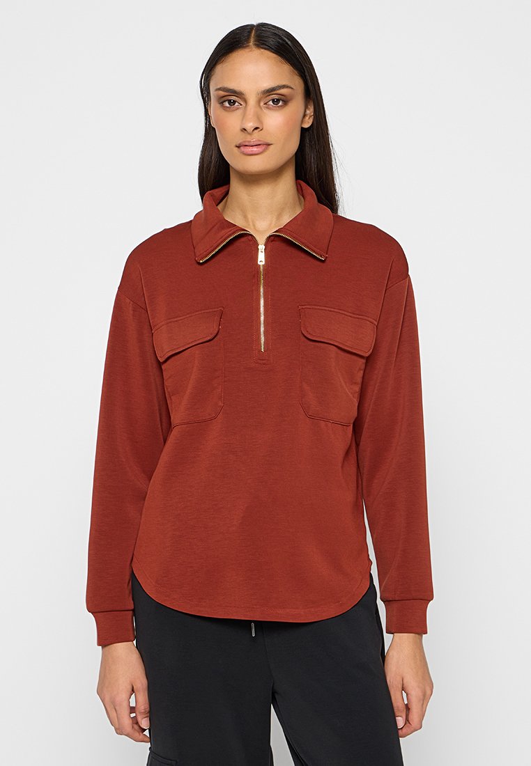 Vrouw in een roestkleurig shirt met lange mouwen, twee borstzakken en een voorrits, gecombineerd met zwarte broek, staande tegen een effen achtergrond.