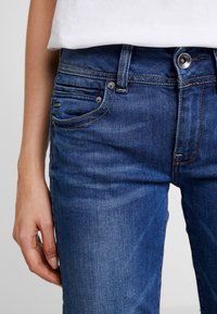 Gros plan sur la main d'une personne reposant à côté d'un jean en denim bleu avec des poches visibles, passants de ceinture et une braguette à boutons, portant une chemise blanche.