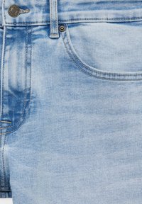 Gros plan sur un jean en denim bleu clair montrant la ceinture, le passant de ceinture, la poche avant, le bouton et les détails des coutures visibles.
