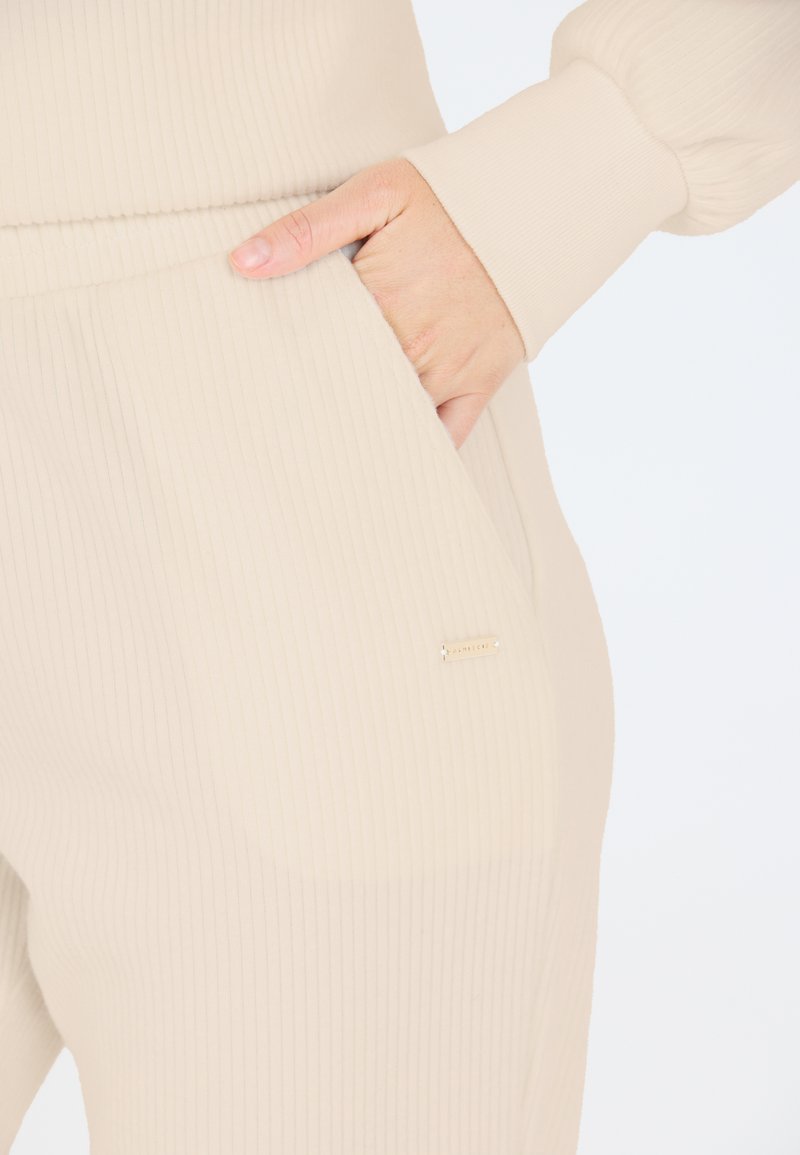 Beige geripptes Stoff-Lounge-Set mit einem dezenten goldenen Markenschild, featuring Seitentaschen und enganliegenden Bündchen. Weiche Textur und komfortables Design.