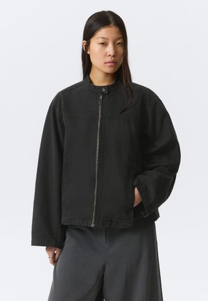 Vrouw met lang donker haar, gekleed in een zwart jack met ritssluiting en drukknopkraag, en losse grijze broek, tegen een effen lichte achtergrond.