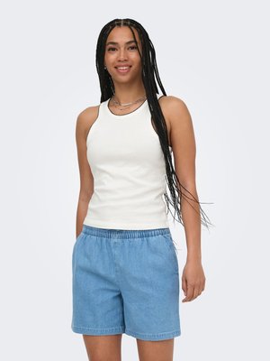 Mujer sonriente con largas trenzas, vestida con una camiseta blanca sin mangas y pantalones cortos de mezclilla azul claro y sueltos, de pie con las manos en los bolsillos.