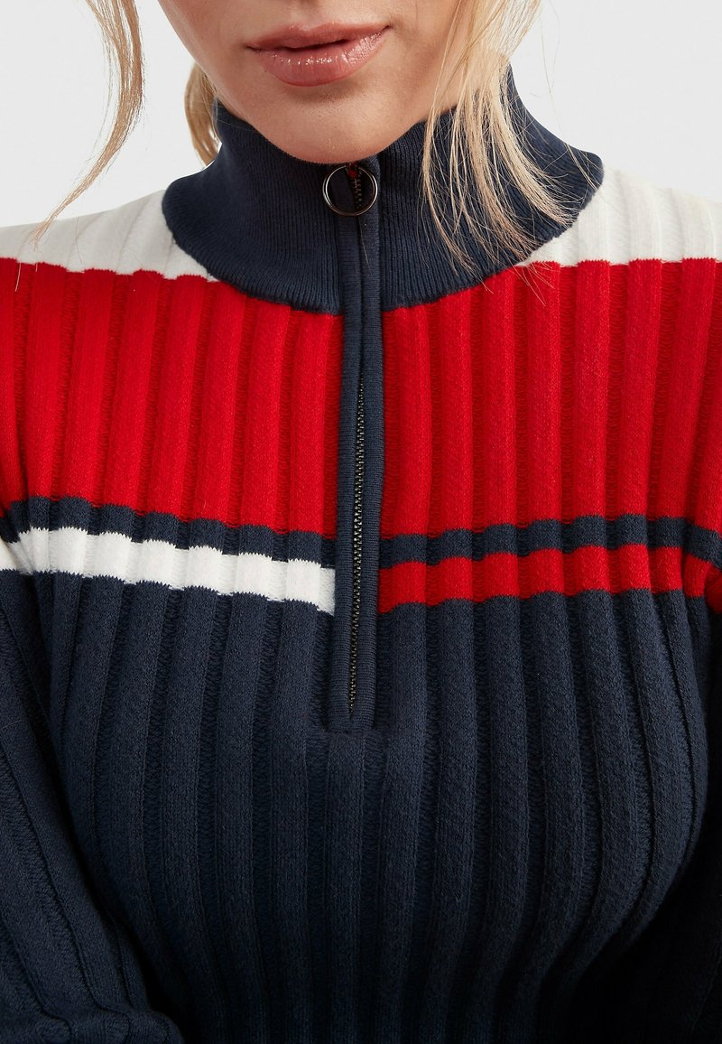 Femme portant un pull en maille côtelée avec un col demi-zip, présentant des rayures horizontales rouges, blanches et bleu marine.