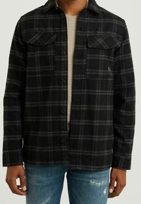 Black and gray plaid flannel shirt with button closure, two chest pockets, and long sleeves, worn over a ribbed light-colored sweater.