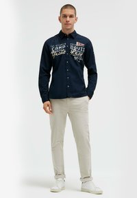 Marineblaue Hemdjacke mit weißen und goldenen Stickereien, die Logos zeigt; kombiniert mit hellbeigen Hosen und weißen Sneakers. Glatte Stofftextur.