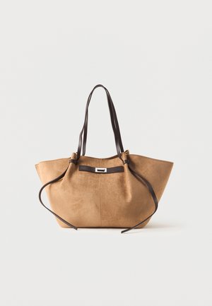 Τσάντα tote από καφέ σκούρα σουέτ με μακριές σκούρες καφέ δερμάτινες λαβές και διακοσμητικό λουράκι μπροστά σε λευκό φόντο.