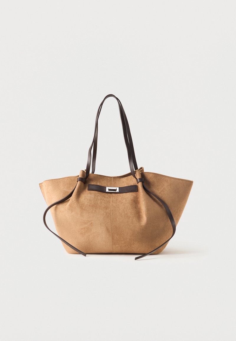 Τσάντα tote από καφέ σκούρα σουέτ με μακριές σκούρες καφέ δερμάτινες λαβές και διακοσμητικό λουράκι μπροστά σε λευκό φόντο.