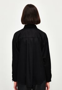 Maglietta nera a maniche lunghe con colletto, caratterizzata da un dettaglio plissettato sulla schiena e polsini con bottone. Texture liscia, design fluido, orlo leggermente curvato.
