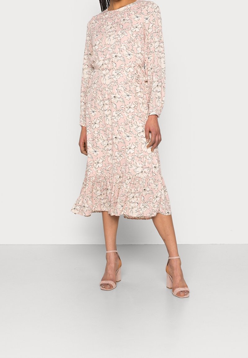 Růžové květinové šaty s dlouhými rukávy, volánkovým lemem a přiléhavým pasem. Design obsahuje černé obrysy květin na jemném pozadí.