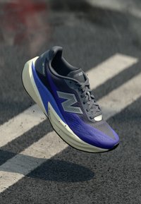 Σκούρο μπλε και γκρι αθλητικό παπούτσι με παχύ λευκό πέλμα και λογότυπο "N", στον αέρα πάνω από άσφαλτο με λευκές λωρίδες διάβασης πεζών.