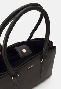 Czarna skórzana torebka z dwoma zaokrąglonymi uchwytami, zapięciem na zatrzask, wewnętrznymi kieszeniami i złotym akcentem z logo. Teksturowana powierzchnia.