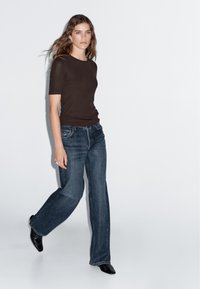 Hnědý krátký rukáv top v kombinaci s tmavě modrými širokými džínami. Model má na sobě černé špičaté boty a stojí na neutrálním pozadí.