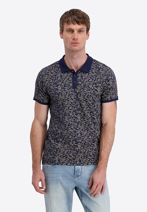 Man wearing navy blue polo shirt with light-colored abstract patterns and light blue jeans, standing against plain background.