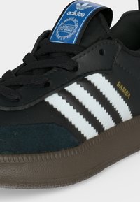 Μαύρο παπούτσι Adidas Samba με λευκές ρίγες, καφέ σόλα, μαύρα κορδόνια, μπλε ετικέτα στη γλώσσα και χρυσό κείμενο "SAMBA" στην πλαϊνή επιφάνεια.