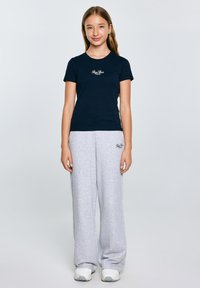 Tamsiai mėlyna trumpomis rankovėmis marškinėliai su logotipu krūtinėje, derinami su šviesiai pilkais sportiniais džinsais, turinčiais logotipą ant klubo, užbaigti baltais sportiniais batais.