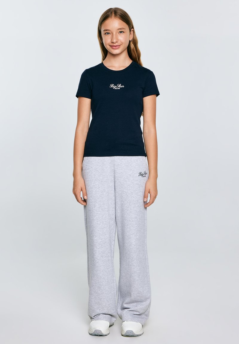 Tamsiai mėlyna trumpomis rankovėmis marškinėliai su logotipu krūtinėje, derinami su šviesiai pilkais sportiniais džinsais, turinčiais logotipą ant klubo, užbaigti baltais sportiniais batais.