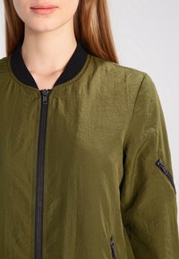 Olijfgroene bomberjack van lichtgewicht materiaal. Heeft een geribde zwarte kraag en twee met ritsen afsluitbare zijzakken. Soepele, matte afwerking.