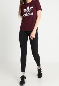 Mujer con una camiseta marón de Adidas, leggings negros, calcetines a rayas en blanco y negro, y zapatillas Adidas negras caminando hacia adelante.