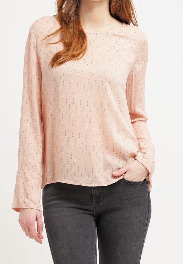 Femme portant un chemisier rose clair à manches longues avec de petites lignes verticales et un jean gris foncé, une main dans la poche.