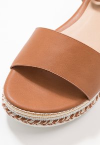 Sandale en cuir brun à glisser avec une large bande et une semelle en espadrille tissée comportant des fils tan et blancs alternés.