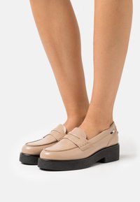 Jambes portant des mocassins en cuir verni beige avec des semelles épaisses noires, présentés sur un fond blanc.