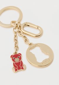 Guld nøglering med et rød emalje-bjørnecharms og et rundt tag med en bjørne-siluet og "FURLA" indgravering. Kædeledsforbindelse inkluderet.