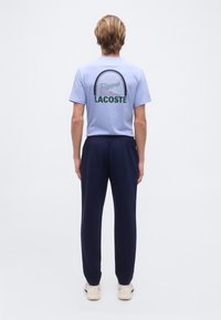 Άτομο που φοράει ένα α νοιχτό γαλάζιο t-shirt Lacoste με γραφική απεικόνιση ρακέτας τένις και σκούρο μπλε παντελόνι, στέκεται με την πλάτη γυρισμένη σε απλό φόντο.