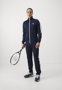Marine trainingspak met witte zijstrepen, ritsjack en bijpassende broek. Zwarte tennisracket in de rechterhand, sportschoenen met rode accenten.