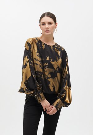 Blouse en satin noir et or tie-dye avec de longues manches volumineuses et des poignets boutonnés, associée à un pantalon noir ajusté.