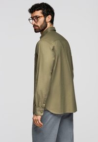 Homme barbu aux cheveux bouclés portant des lunettes écaille de tortue, une chemise à manches longues vert olive et un pantalon gris, regardant par-dessus son épaule.