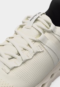 Κοντινό πλάνο σε αθλητικό παπούτσι κρεμ χρώματος με υφασμάτινο άνω μέρος πλεκτό με υφή, κορδόνια από ύφασμα και λογότυπο Columbia στην ετικέτα της γλώσσας.